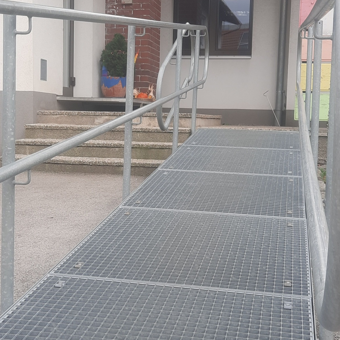 Man sieht den Zugang zu einem Kindergarten, der über eine Rampe führt – welche allerdings vor drei Treppenstufen endet. Hier wird nicht nur Kindern im Rollstuhl der Zugang verwehrt, sondern auch Eltern, die einen Rollstuhl nutzen. Eine Teilnahme an Elternabenden, ein Gespräch mit der Kindergartenpädagogin oder auch der Besuch einer Veranstaltung wird damit unmöglich gemacht.