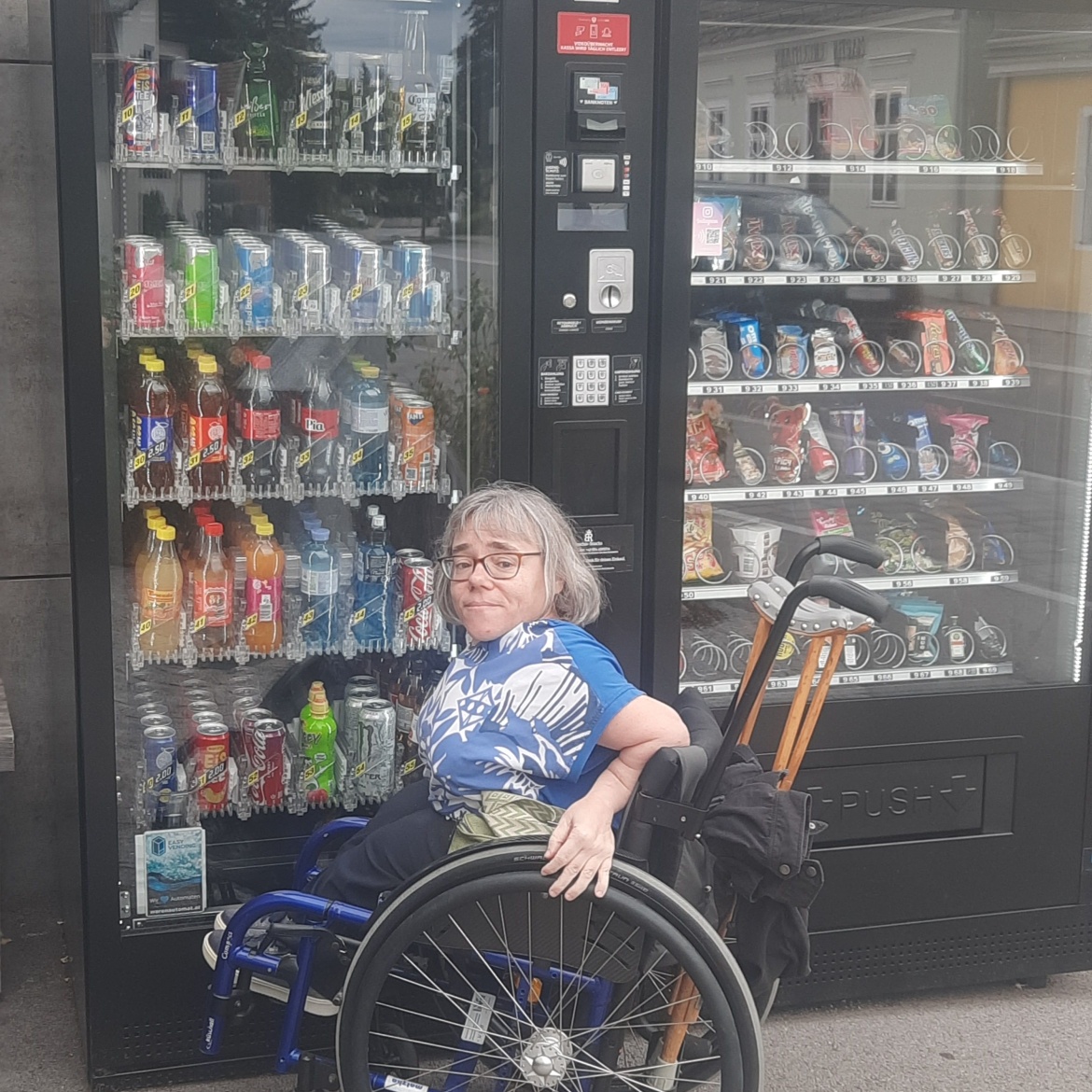 Bildbeschreibung: Sandra Hermann im Rollstuhl vor einem herkömmlichen Snack- bzw. Getränke-Automaten. Die Bedienungselemente des Automaten liegen außerhalb ihrer Erreichbarkeit.