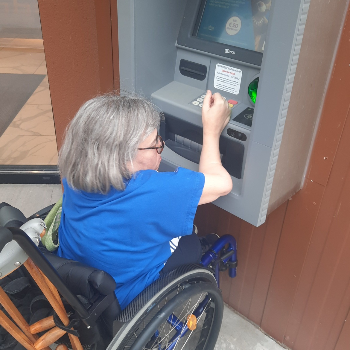 Sandra Hermann im Rollstuhl vor einem herkömmlichen Bankomaten. Das Display ist zu hoch angebracht und kann nicht eingesehen werden. Auch die Bedienungselemente sind nur schwer erreich- bzw. einsehbar.